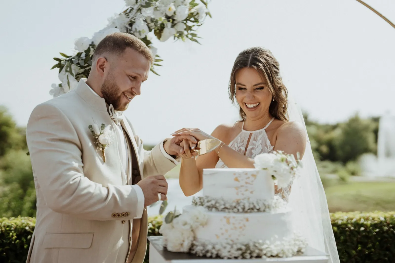 Anna & Moritz – Ein Brautpaar schneidet gemeinsam lachend die Hochzeitstorte im Freien an