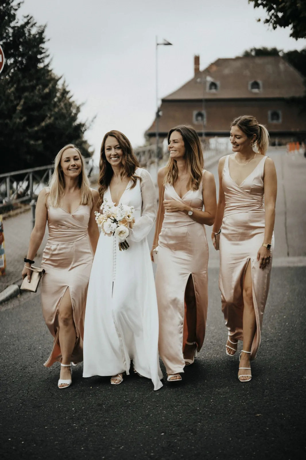 Deana & Sebastian – Eine Braut läuft lachend mit ihren drei Brautjungfern auf einer Straße im Freien