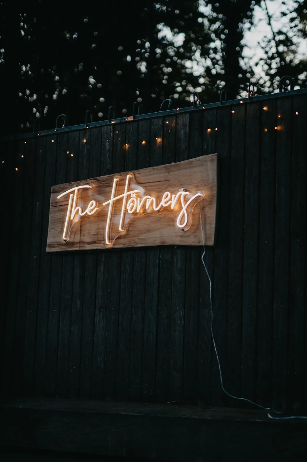 Jana & Christoph – Ein hölzernes Schild mit einem leuchtenden Neon-Schriftzug 'The Horners' an einem Holzzaun