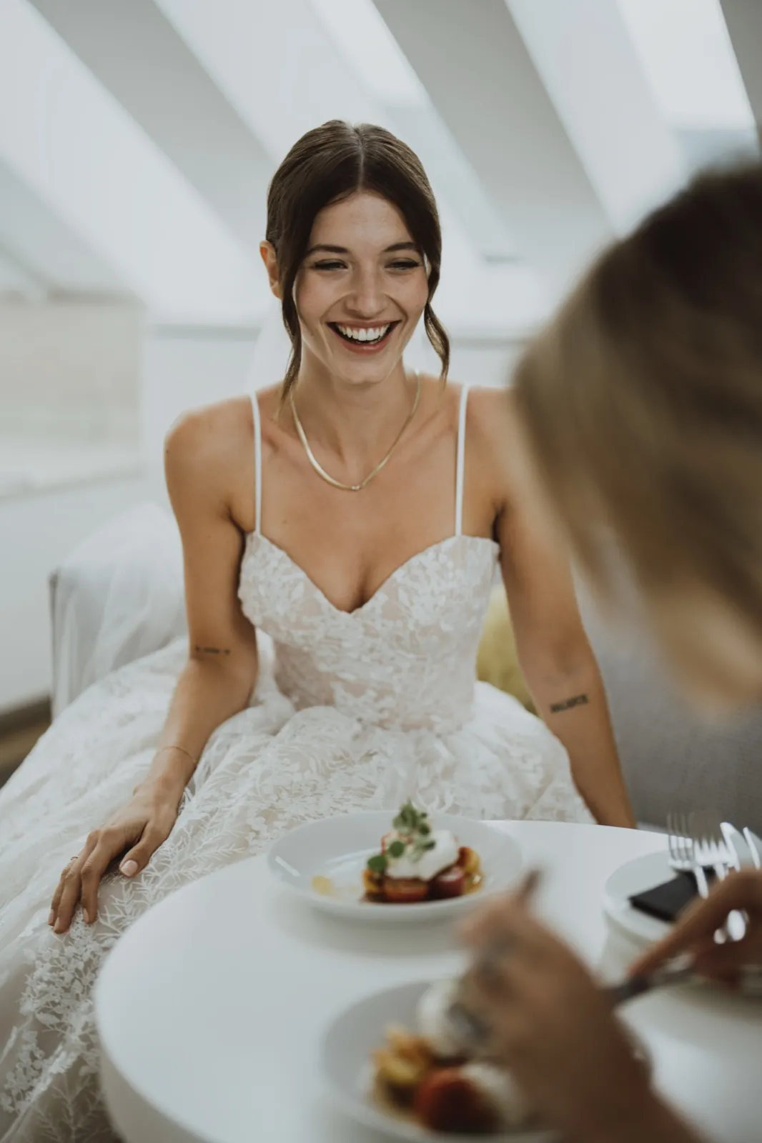 Julia & Chris – Eine lächelnde Braut im weißen Spitzenkleid sitzt an einem weißen Tisch mit Essen