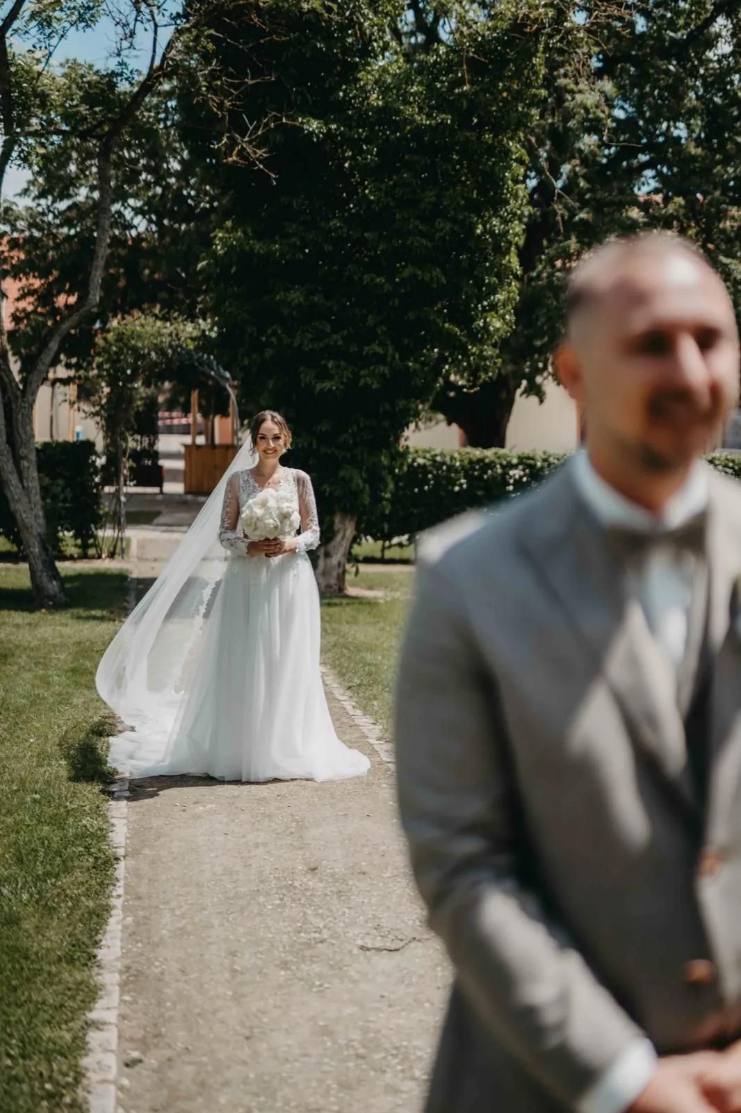 Kristina & Pascal – Eine Braut nähert sich im Freien ihrem im Vordergrund unscharf stehenden Bräutigam